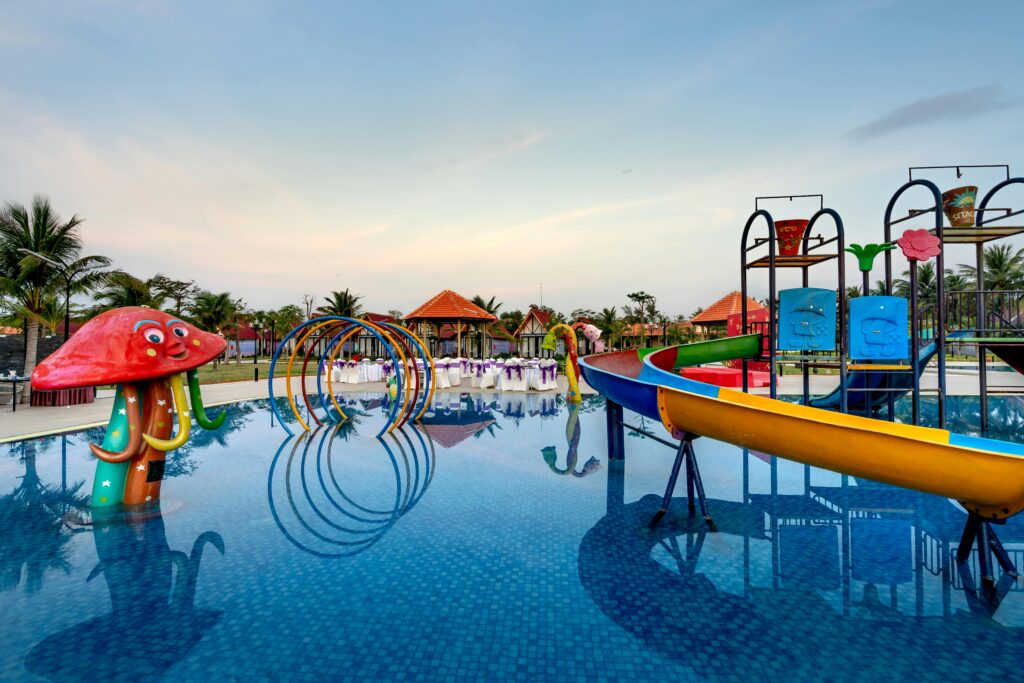 Colorful waterpark with slides, mushroom-shaped feature, and palm trees reflecting in pool.